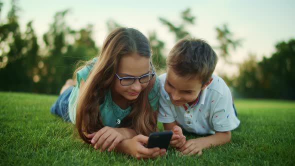 Boy and Girl Lying on Grass with Cellphone. Brother and Sister Using Smartphone alt