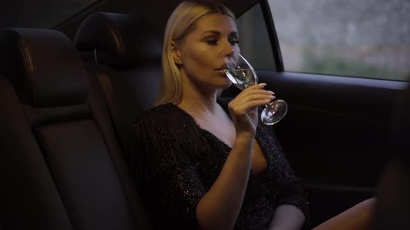 Portrait of Stylish Young Blond Woman Drinking Champagne, Putting Glass Away, and Looking Out Car alt