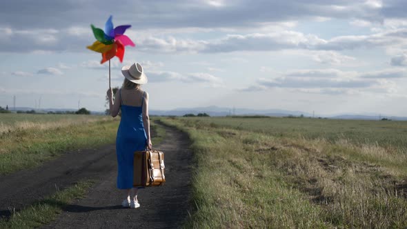 Girl in blue dress with suitcase and pinwheel on country road in summer.