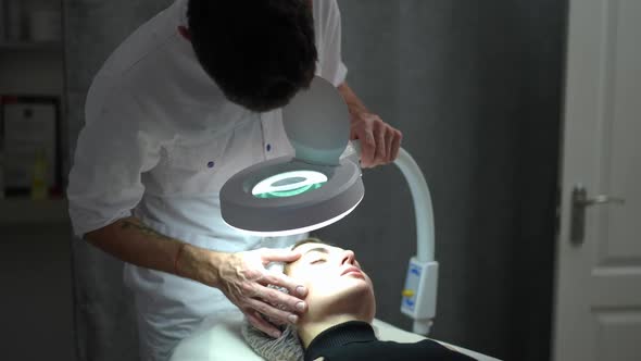 Focused Expert Male Beautician with Magnifying Lamp Examining Face of Female Client Indoors alt