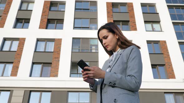 Woman in a Business Suit Looks at the Phone and Rejoices on the Background of a Building on a Sunny alt