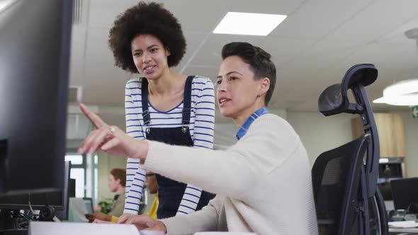 Diverse smiling female business colleagues pointing at computer and discussing in office alt