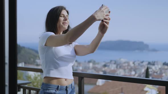 Attractive Millennial brunette  woman making video on cell phone while standing on the terrace alt