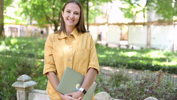 Young Smiling Woman Holding Laptop and Looks at the Camera Standing Outdoors