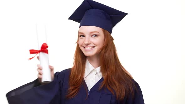 Smiling Ginger Young Graduate Girl in Blue Robe and Square Academical Cap Entering Frame Holding Her alt