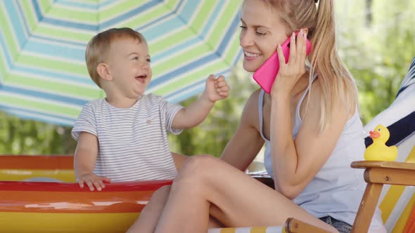 Happy smiling mother with her little baby son, together talking at mobile phone on a sunny summer da