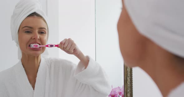 Woman in bathrobe brushing teeth while looking in the mirror alt