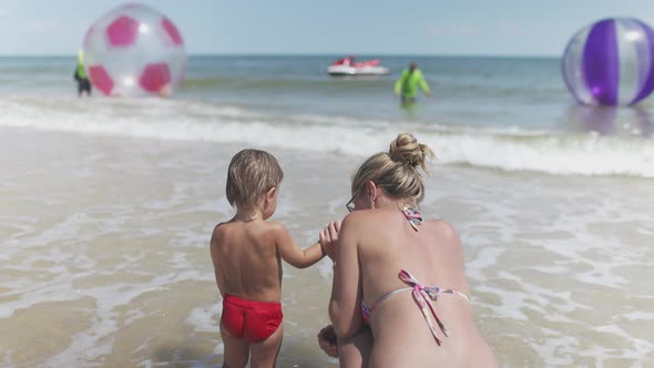 The Kid Looks at the Horizon of the Sea and Inflatable Big Balls with His Mother in a Bikini alt