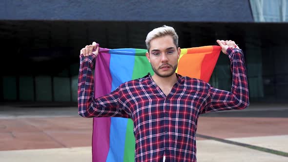 Young gay man holding rainbow flag outdoor at pride parade while smiling on camera