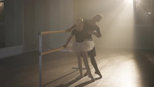 Smiling Slim Ballet Teacher Rehearsing Tendu with Student at Barre in Backlit Fog alt