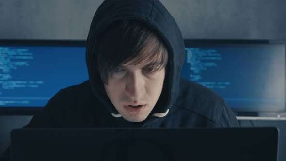 Man in Hood Sitting at Table and Coding on Laptop alt