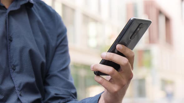 A Man Looks at a Smartphone in an Urban Area  Closeup  Office Buildings in the Blurry Background