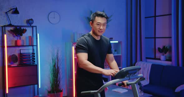 Sporty Asian Guy in Training Clothes Setting the Speed on Treadmill's Control Panel During Evening alt
