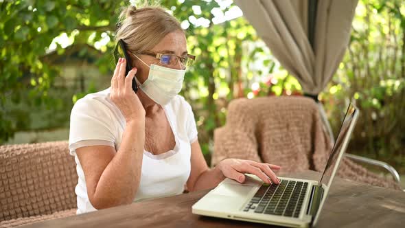 senior woman in protective face mask speaking smartphone working online with laptop computer outdoor