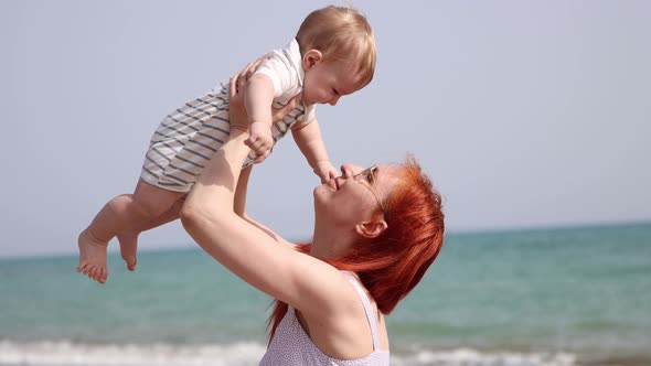 Happy Mother Raising Up Her Little Son Sitting on the Beach alt