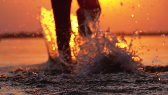 Silhouette of Legs of Boy Running Into the Water at Sunset and Creating Splashes alt
