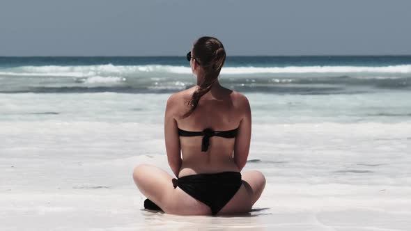 Young Woman in Black Bikini Tans in Lotus Pose on Paradise Sandy Beach By Ocean alt