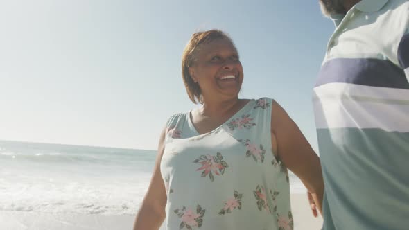 Smiling senior african american couple embracing and walking on sunny beach alt