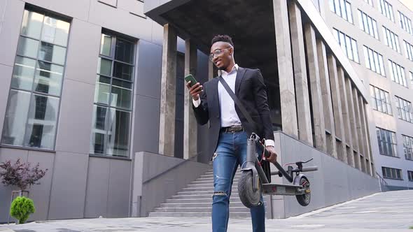 African American in Trendy Wear Walking  with Personal E-Scooter in Hand and Talking on Phone alt