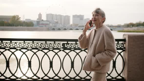 Side View of Senior Lady Chatting on Cellphone Walking in City Crossing Bridge on Summer Day alt