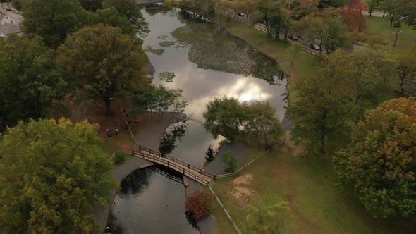 An aerial drone shot over a park in the day with a pond, with an arched footbridge. It's a beautiful alt