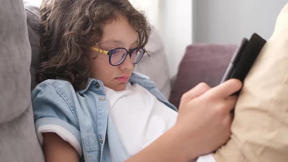 Little Cute Boy with Eyeglasses Sitting on Sofa and Playing Game on Digital Tablet Closeup Portrait alt