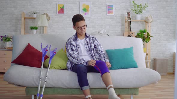 Young Asian Man Sitting on the Couch Puts a Tightening Elastic Bandage on a Sick Knee alt