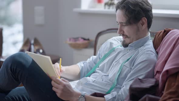 Side View of Smiling Skilled Fashion Designer Drawing with Pencil Sitting on Couch in Atelier alt