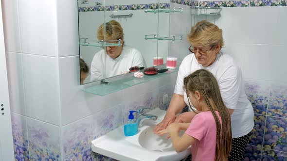 Grandmother teaches a little girl how to wash their hands with soap over a sink