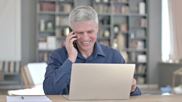Middle Aged Man Talking on Smartphone While Working on Laptop in Office alt