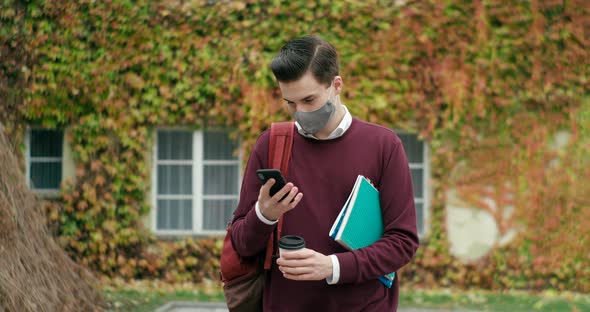Male Student in Mask Using Smartphone Mobile App and Walking Outside with Coffee