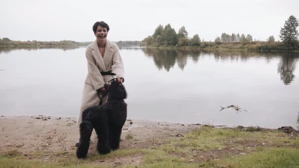 An Elegant Young Brunette in a White Coat Stands By the River and Holds a Large Black Poodle on a alt