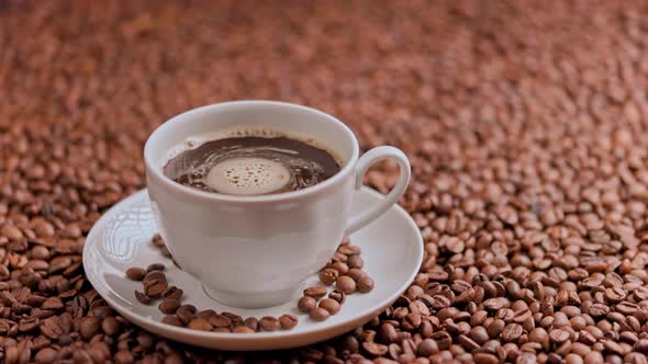 Black Coffee in a White Cup with Spinning Bubbles on a Flat Surface Covered with Roasted Coffee