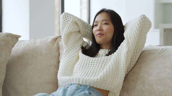 Beautiful Young Asian Woman in a Sweatshirt Resting on the Sofa in the Living Room Throws Her Hands alt