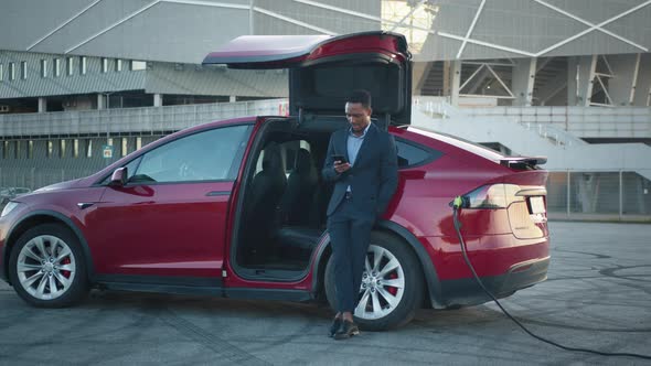 African American Male Leaning on Luxury Red Auto and Looking at Mobile Screen alt