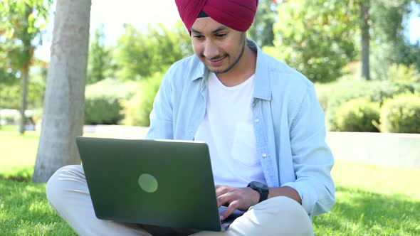 Male Indian Student Wearing Red Traditional National Turban Using Laptop Computer Sitting on Lawn