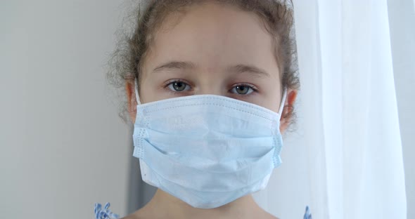 A Child at Home in a Protective Medical Mask with a Sad Face and Eyes Looks at the Camera alt