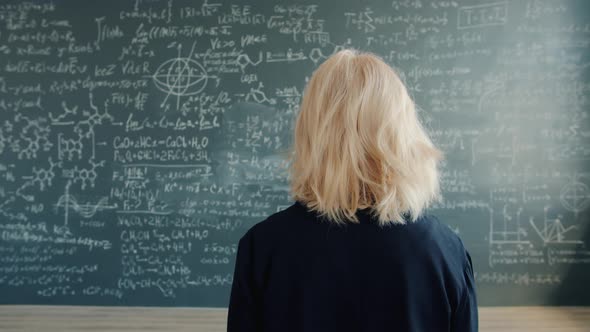 Female Scientist Walking To Chalkboard Wall with Formulas in University Classroom alt
