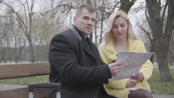 Cheerful Caucasian Married Couple Talking and Looking at Map As Sitting in Park with Travel Bag alt