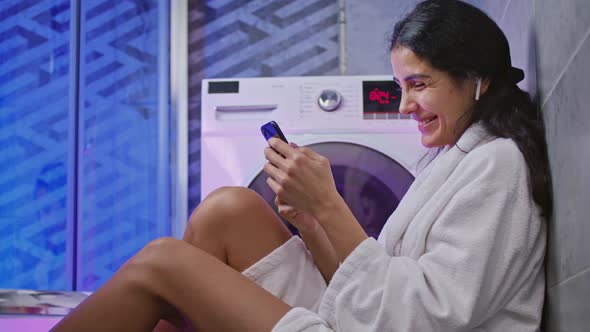 Mixedraces Girl Sitting on Floor and Chatting on Smartphone While Washing Machines Working