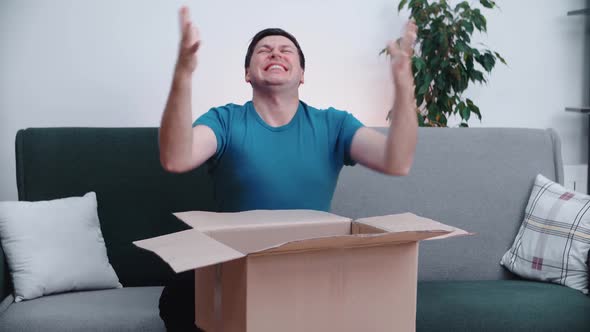 Young man opens a cardboard box while sitting on a home office couch alt