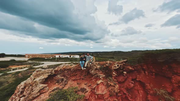 Zooming out shot by drone featuring two men sitting on top of the cliffs and hugging alt