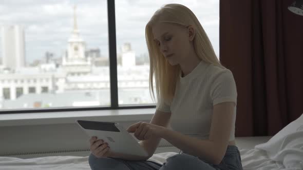 A Young Blonde Woman Works Behind a Laptop Near a Large Window alt