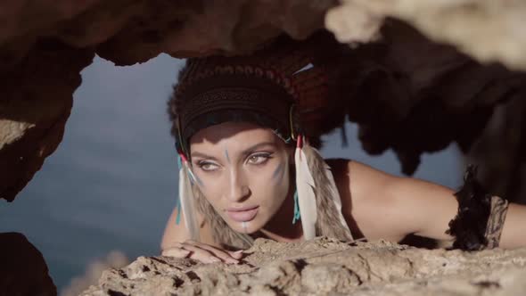 Strong Free Wild Woman with Tribal Ornamental Makeup and Feather Hat Hiding in Rocks and Hunting alt