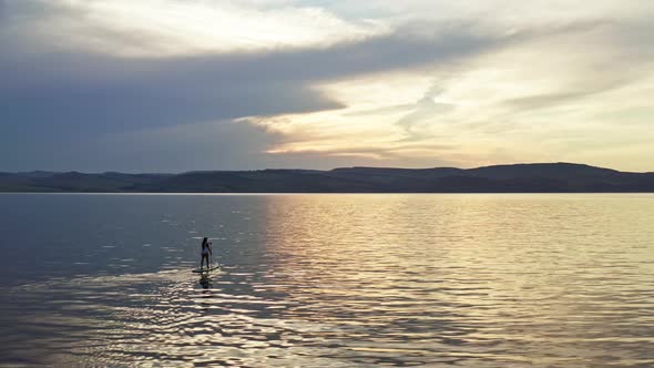 Drone Shot of a Silhouette of a Young Woman Floating on a Board with a Paddle on a Mountain Lake at