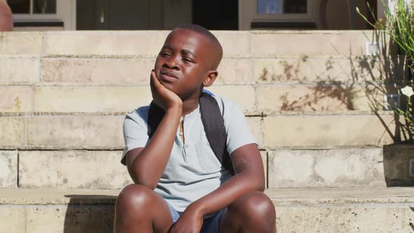 Sad african american boy wearing face mask sitting on stairs outdoors on a sunny day alt