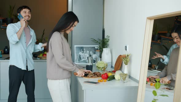 Young Woman Cooking Food While Loving Husband Chatting on Mobile Phone and Kissing Wife in Kitchen alt