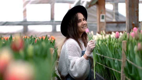 Happy Young Farmer Woman in a Hat Sings a Song with a Tulip Flower While Working in a Greenhouse alt