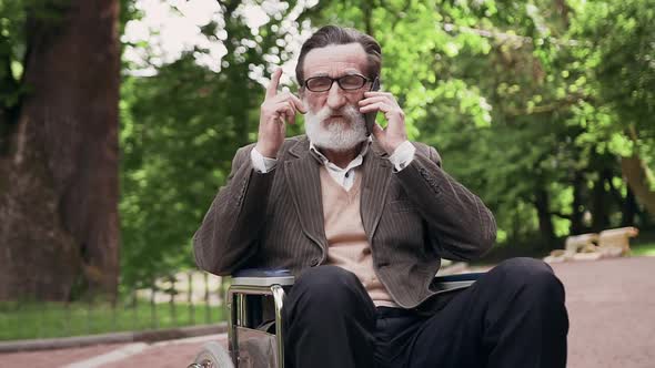 Old Bearded Man in Glasses Sitting in Wheelchair in the Urban Park and Talking on Mobile alt