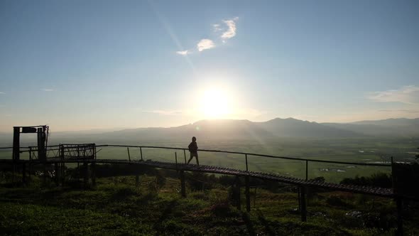 Slow motion silhouette of a woman walking on bamboo wooden bridge with mountain views and sunset sky